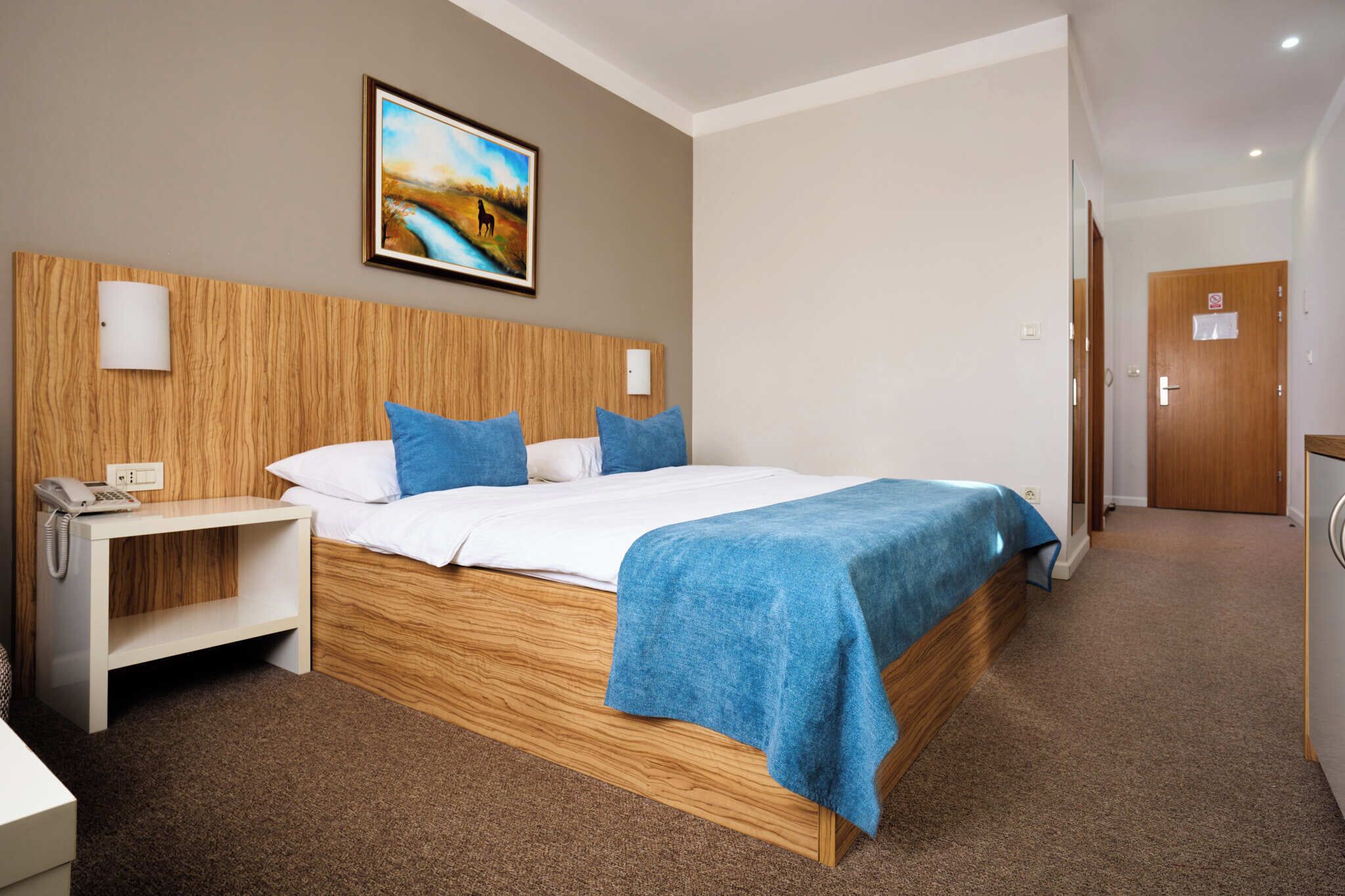 Habitación de hotel con cabecera de madera, cama con ropa de cama azul, mesita de noche con un teléfono y un cuadro en la pared.