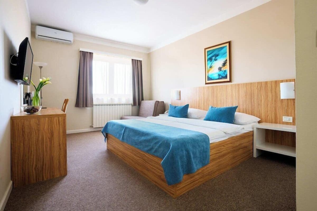 A hotel room with a bed, a desk, a TV, a window with curtains, and a painting on the wall.