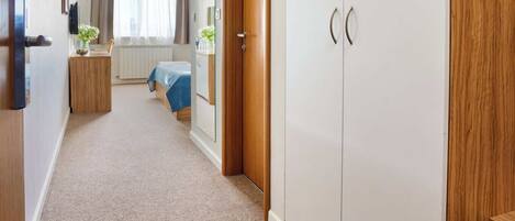 A hallway with a carpeted floor, a wooden wardrobe, a bed with a blue cover, and a small table with a plant.