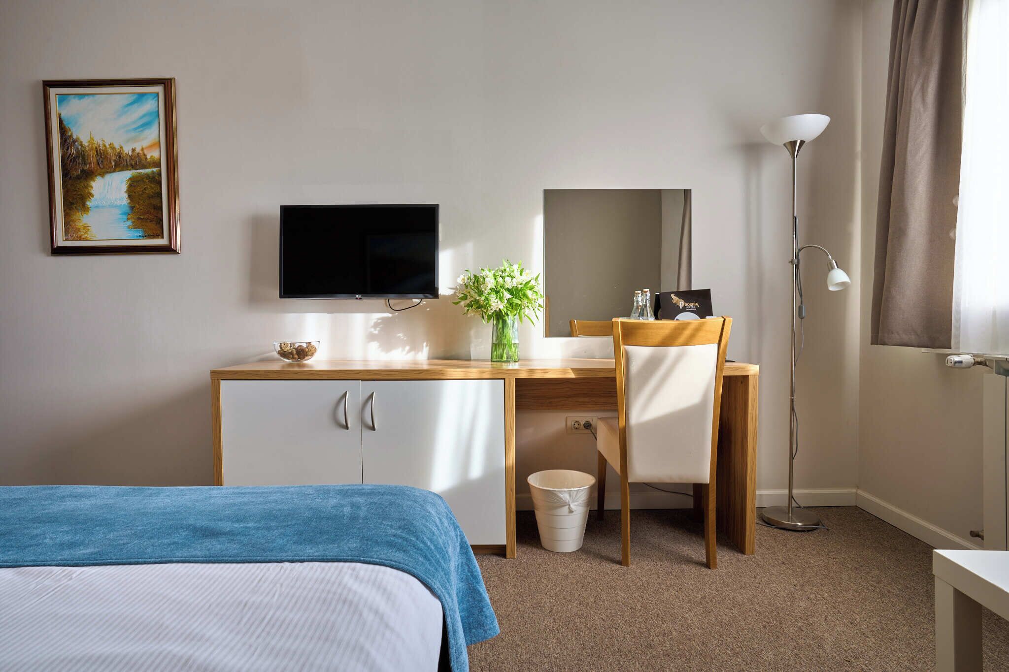 A hotel room with a bed, a desk with a chair, a television, a vase with flowers, a floor lamp, and a painting on the wall.