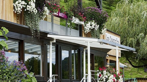 Un restaurante con un letrero, mesas al aire libre con cestas de flores colgantes y vistas a zonas verdes al fondo.