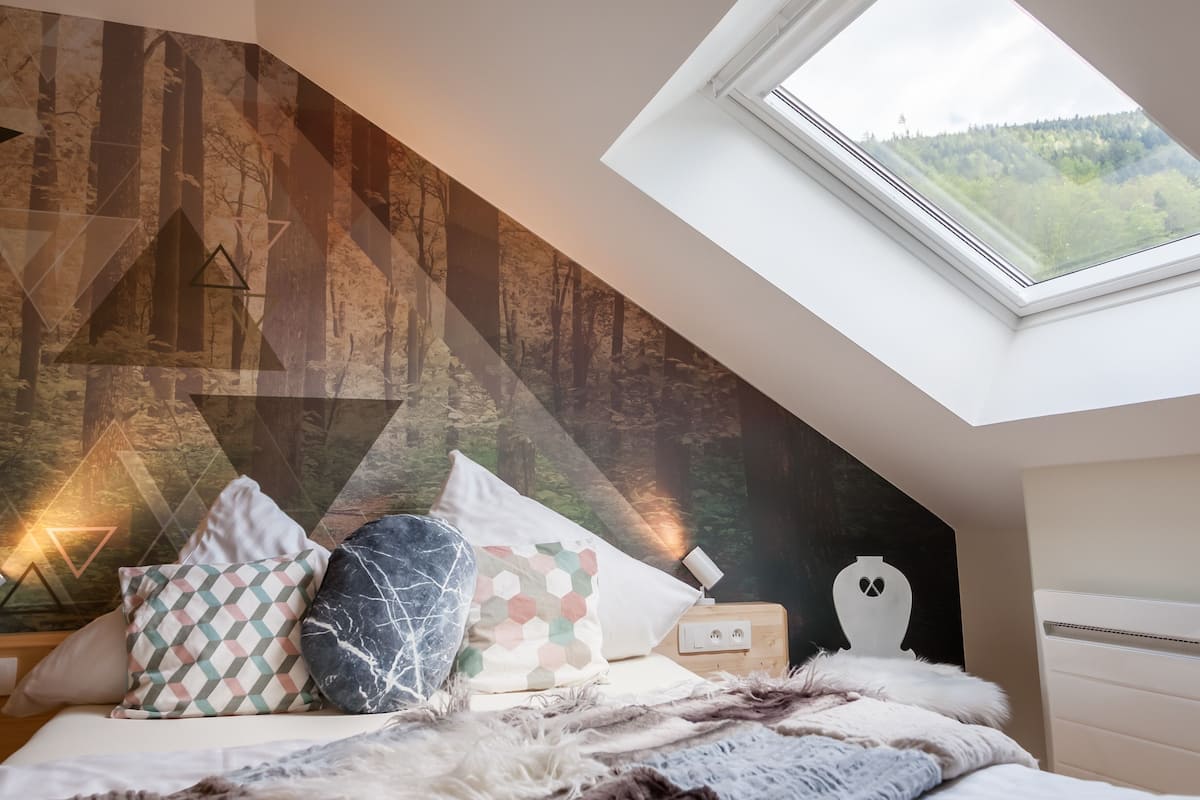 A bedroom with a sloped ceiling, a skylight, a bed with patterned pillows, a wooden bedside table, and a wall with a geometric design.