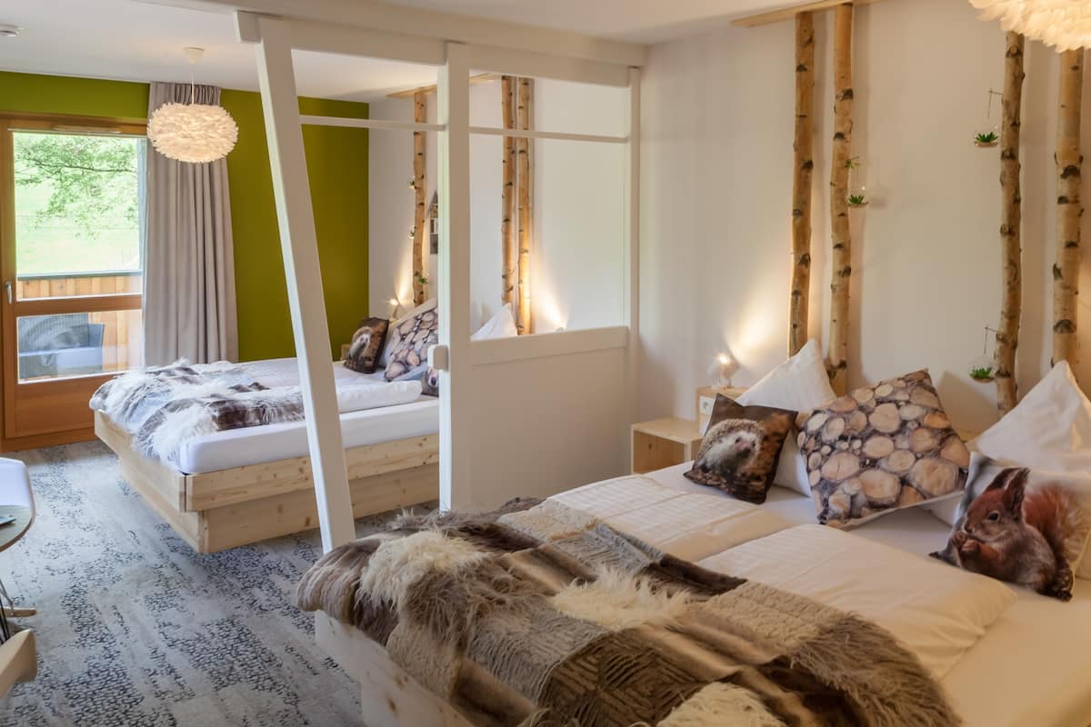 A cozy bedroom with a large bed, a smaller bed, and a window with curtains. There are pillows and a blanket on the beds.