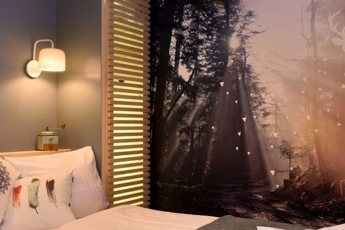 A bedroom with a bed, a nightstand, a wall-mounted lamp, and a forest-themed tapestry.