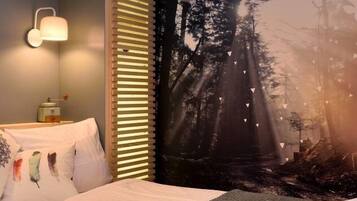 A bedroom with a bed, a nightstand, a wall-mounted lamp, and a forest-themed tapestry.