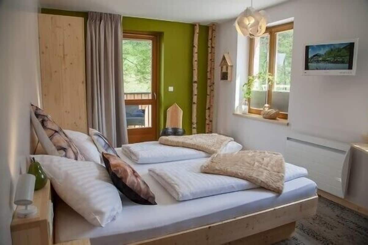 A double bed with pillows, a wooden headboard, a small table with a lamp, a window with a view, and a painting on the wall.
