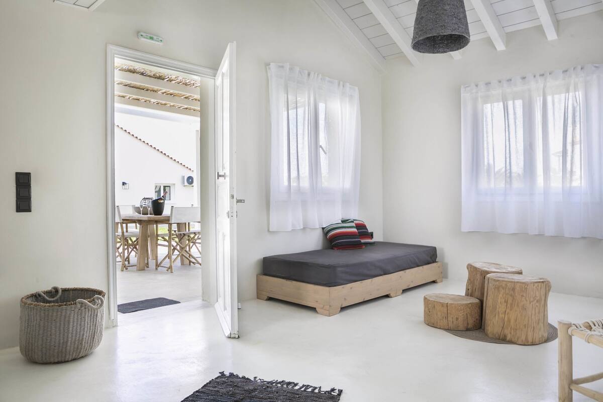 Ein schlicht eingerichtetes Schlafzimmer mit einem Bett, einem kleinen Tisch und einem Fenster mit durchsichtigen Vorhängen.