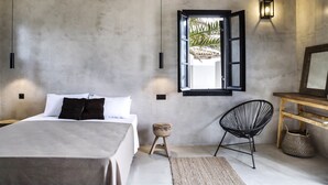 Een slaapkamer met een bed, een plafondventilator, een raam met open luiken, een wandlamp, een stoel en een klein krukje.