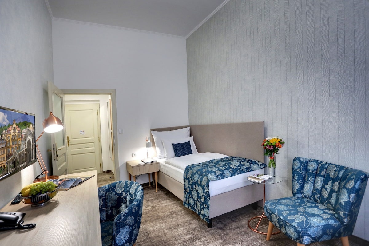 A hotel room with a bed, a desk with a lamp, a chair, a small table with a vase of flowers, and a painting on the wall.