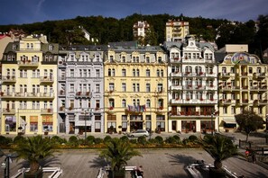 A row of historic buildings with balconies and flags, a paved plaza with palm trees, and a car parked in front. - ASTORIA Hotel & Medical Spa (Karlovy Vary)