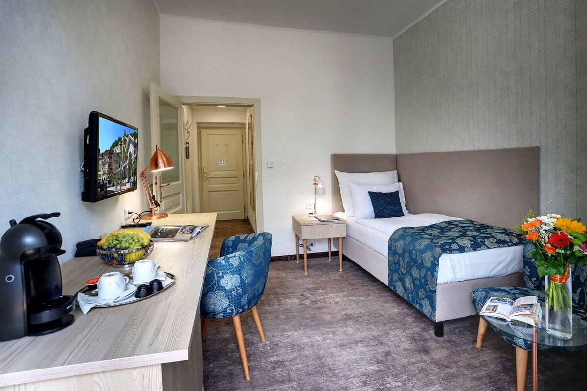 A hotel room with a bed, a desk, a chair, a coffee maker, a TV, and a door leading to another room.