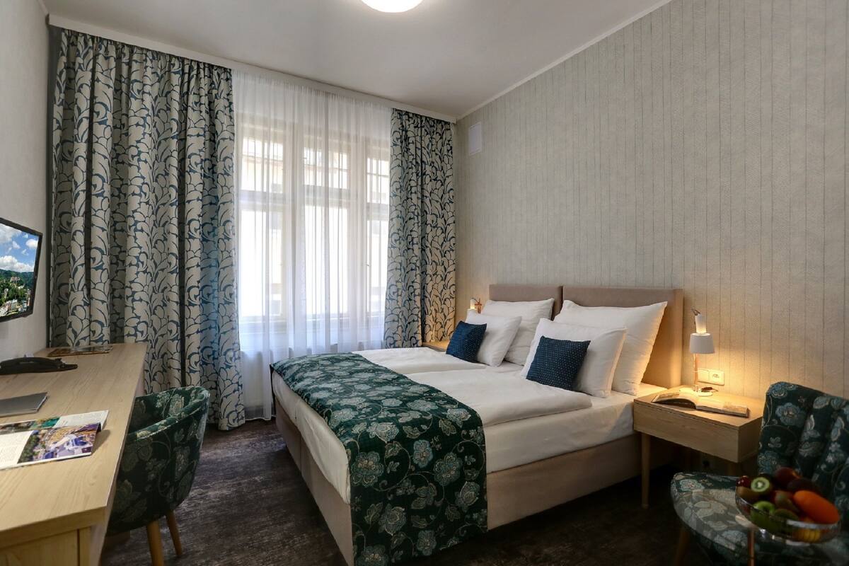 A hotel room with a bed, bedside table, lamp, chair, desk, and a fruit bowl.