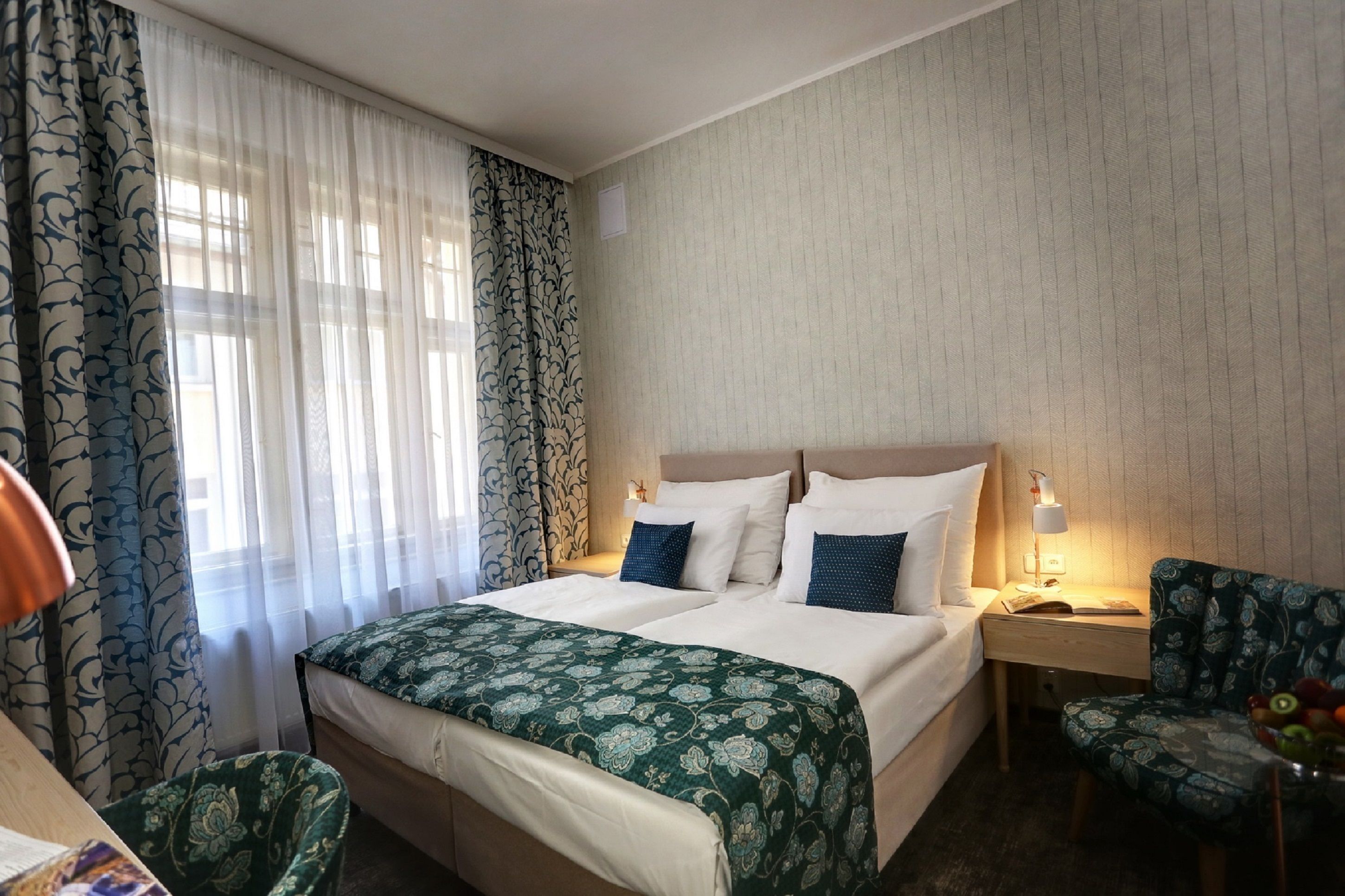 Una habitación de hotel con cama, mesita de noche, silla y ventana con cortinas.