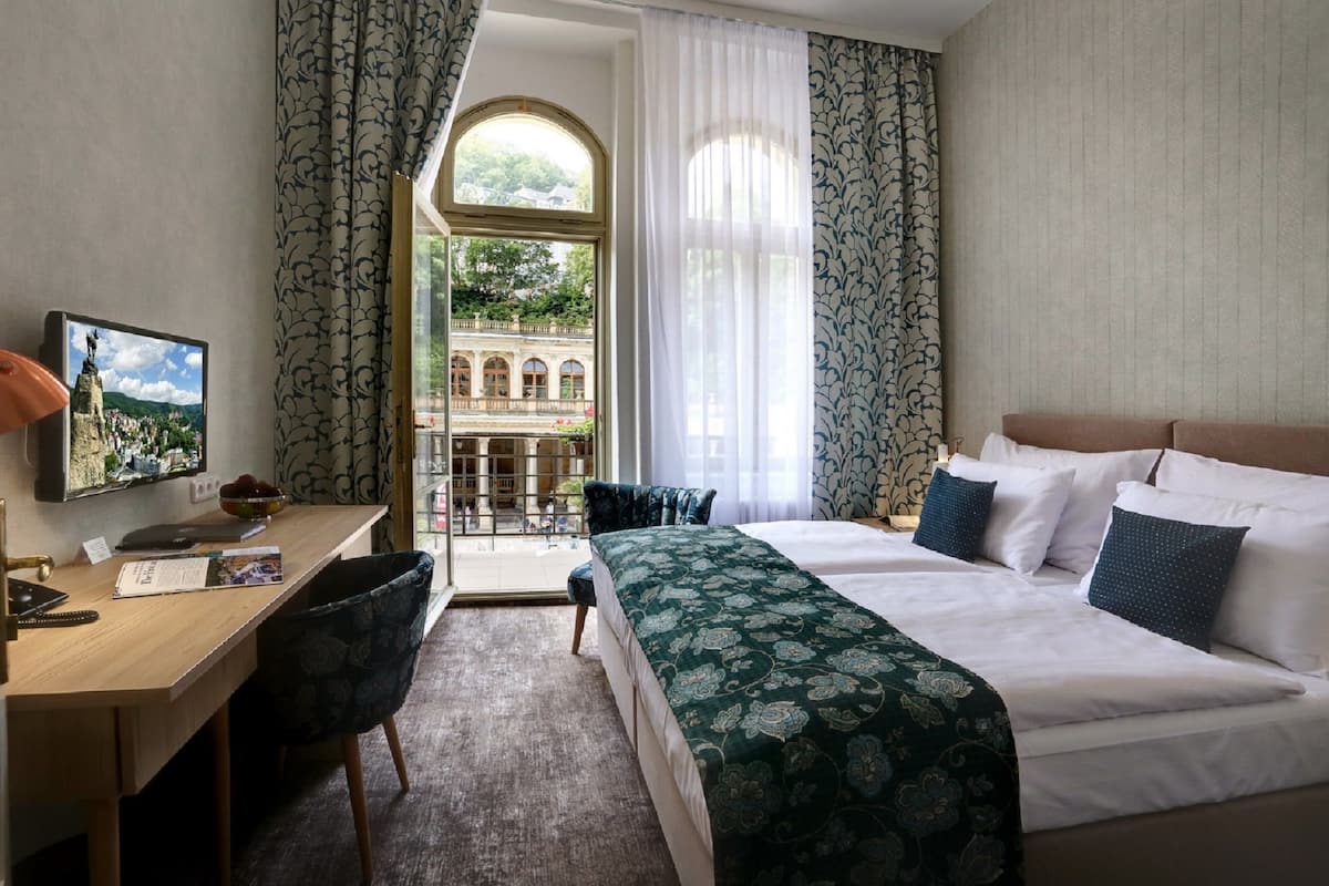 A hotel room with a bed, desk, chair, and a view of a courtyard through a window.
