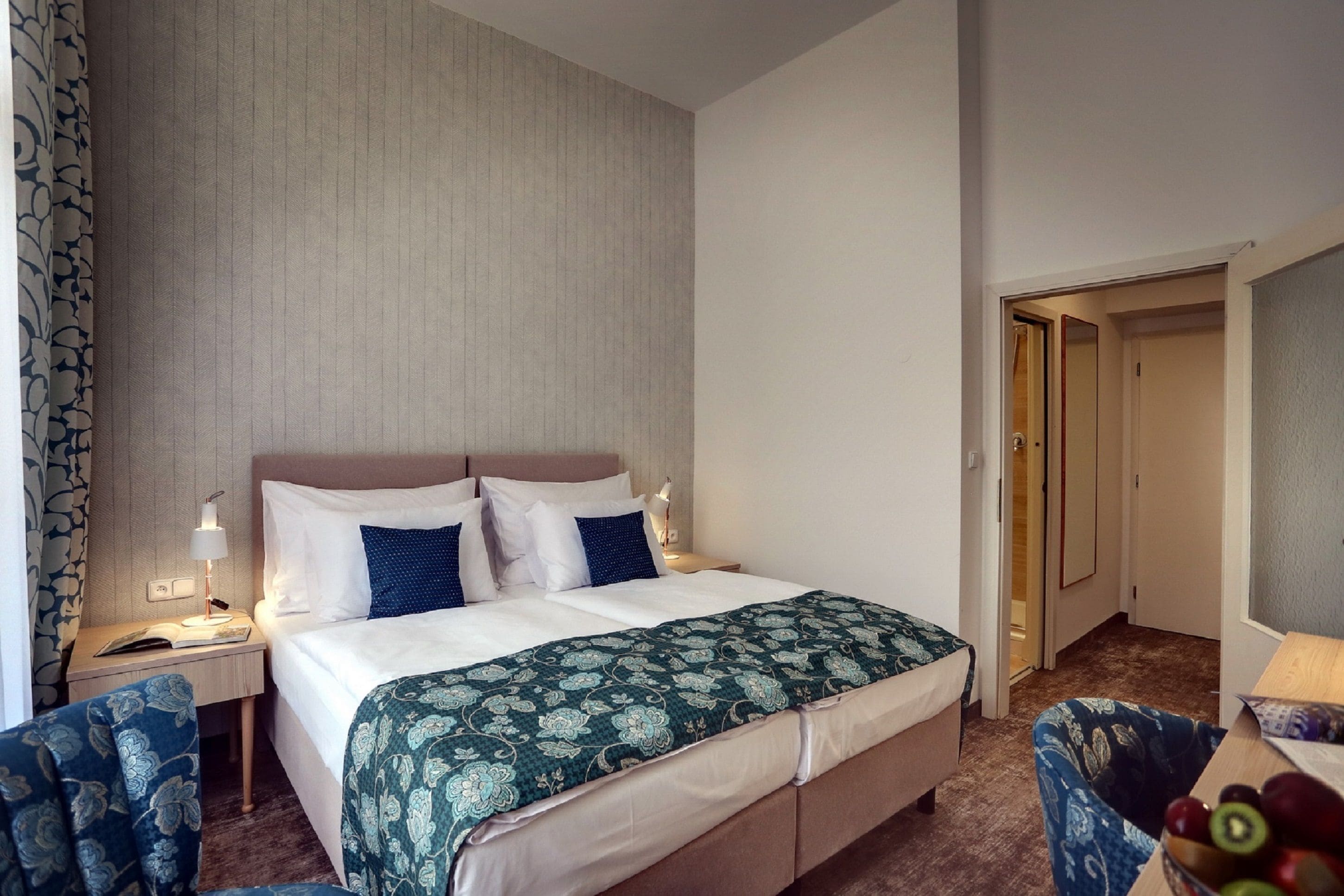 A hotel room with a bed, bedside tables, a chair, and a door leading to another room.