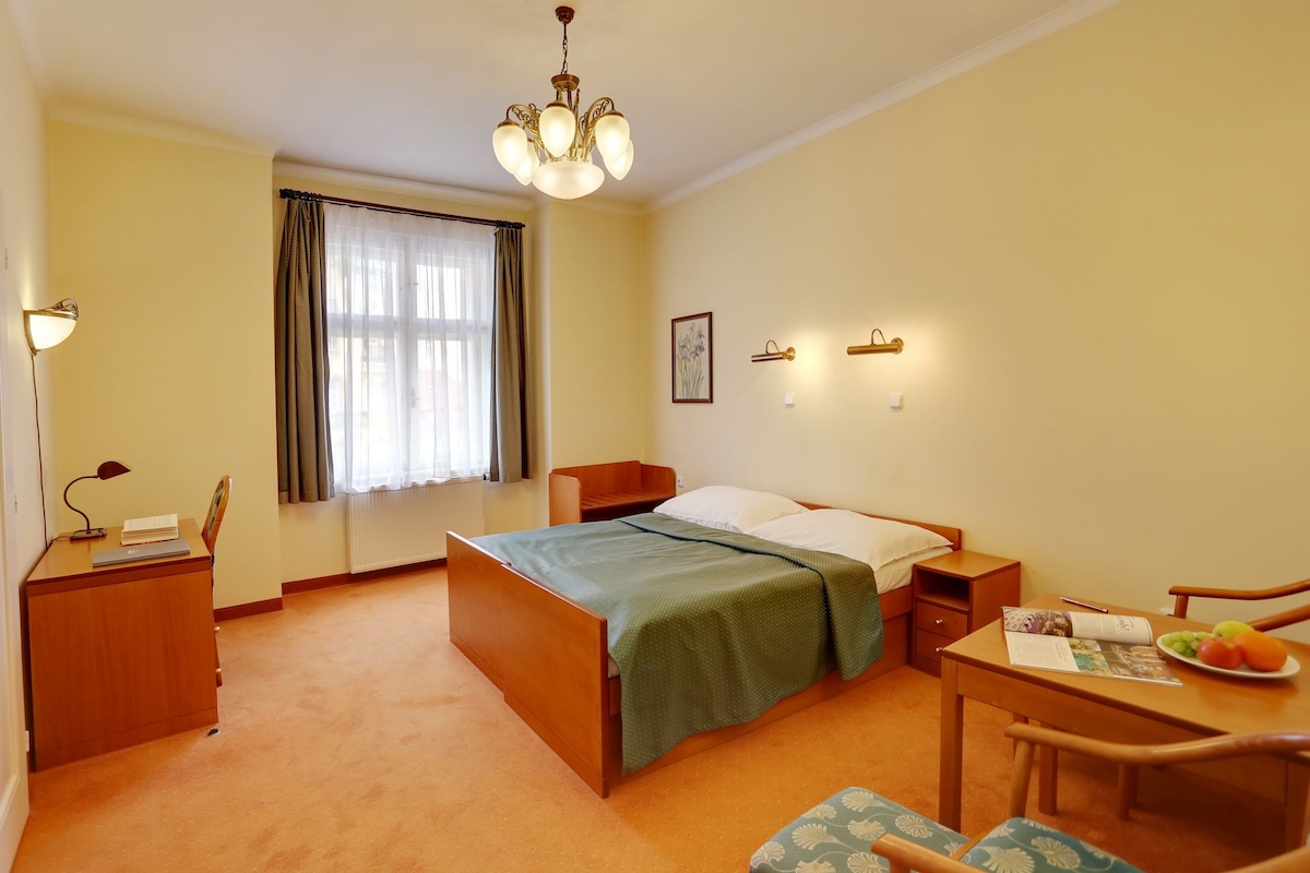 A hotel room with a bed, bedside tables, a desk with a chair, a dining table with a chair, a chandelier, and a window with curtains.