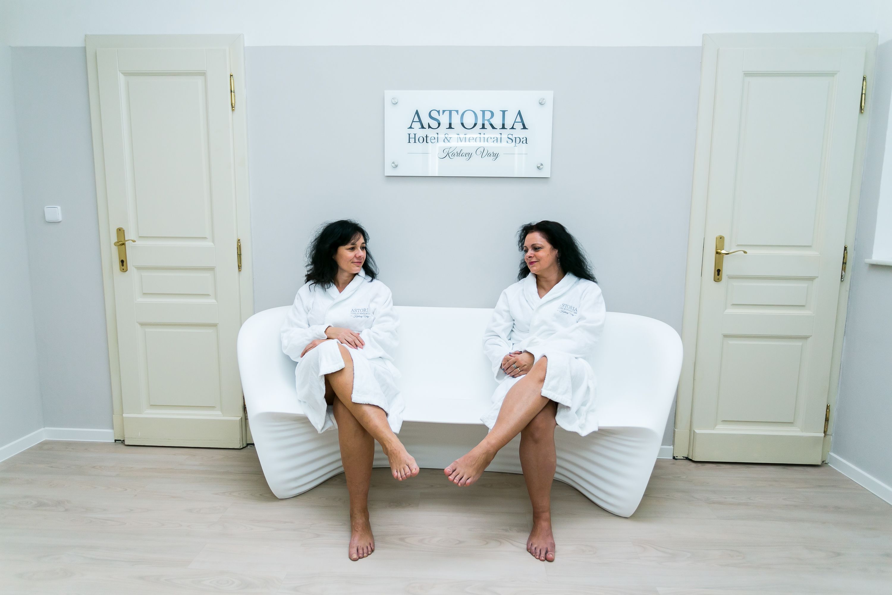 Two individuals seated on a white couch in a room with light-colored walls and wooden flooring. A sign above them reads "ASTORIA Hotel & medical Spa."