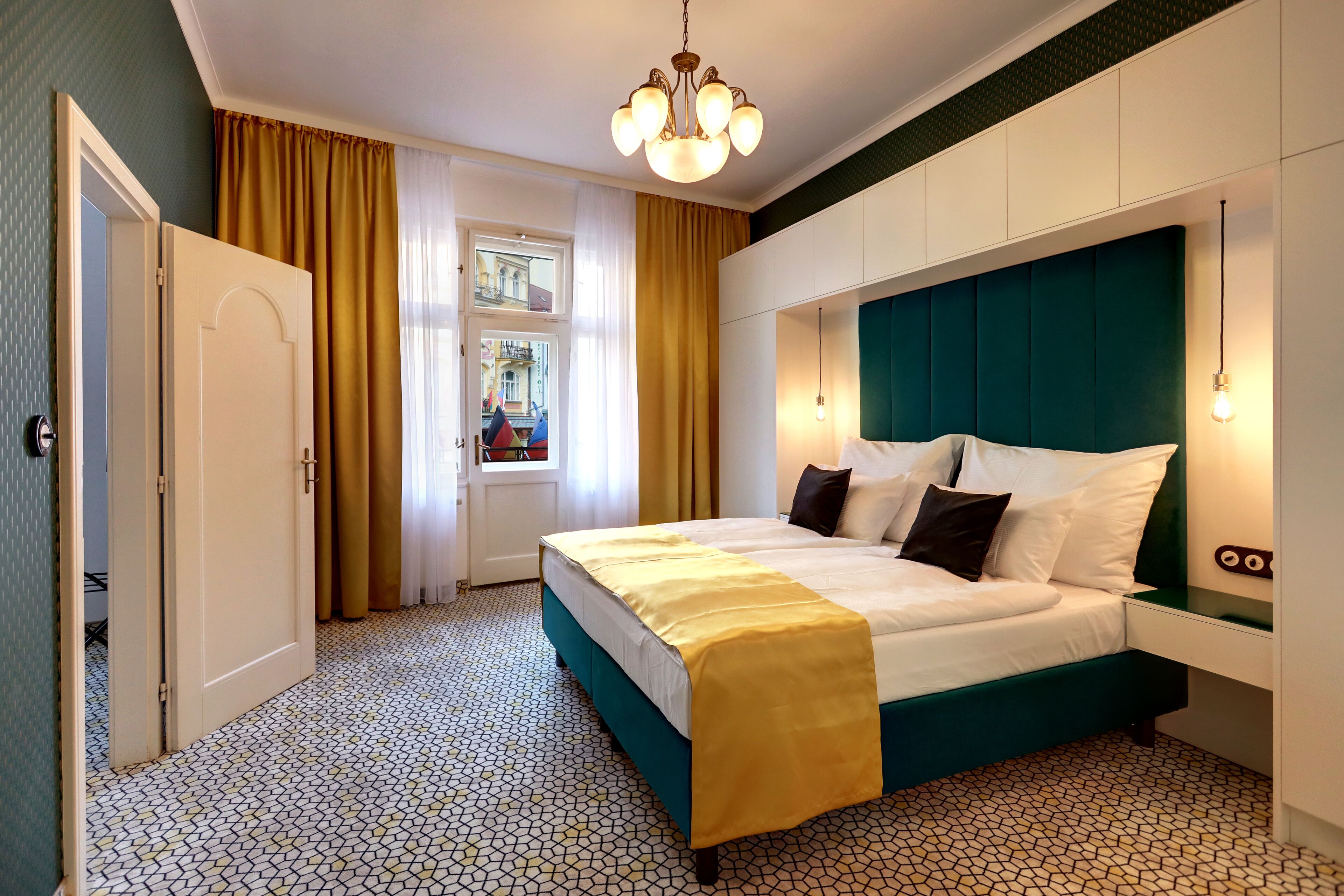 Habitación de hotel con cama, ventana con cortinas, candelabro y piso de baldosas.