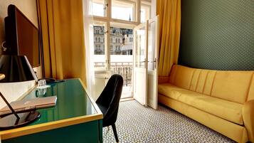 Una habitación con un escritorio verde, una silla negra, un sofá amarillo y vistas a edificios a través de una ventana.