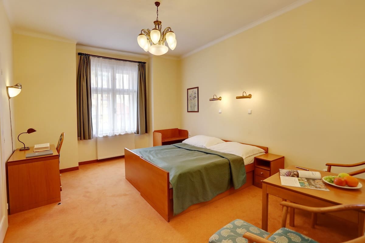 Ein Hotelzimmer mit Bett, Nachttischen, einem Schreibtisch mit Stuhl, einem Esstisch mit Stuhl, einer Chandelierleuchtre und einem fenster mit Vorhängen.