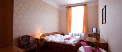 Habitación de hotel con cama, una silla, escritorio, mesa, televisor y ventana con cortinas.