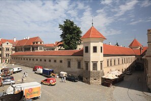 A historic building with a red-tiled roof, a central tower, and a paved courtyard with parked vehicles and people.