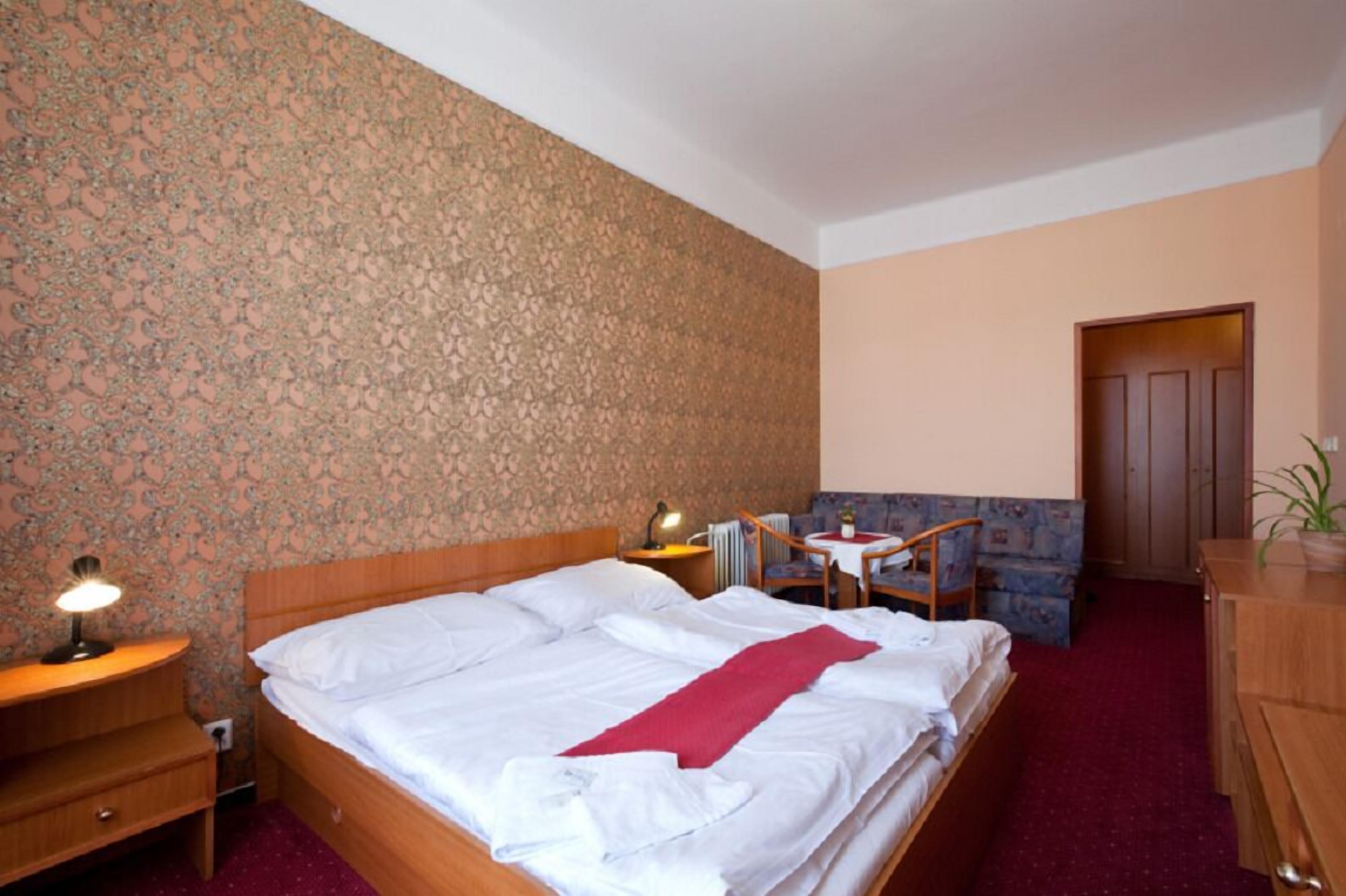 A hotel room with a bed, bedside table, lamp, chair, small table, and a door.