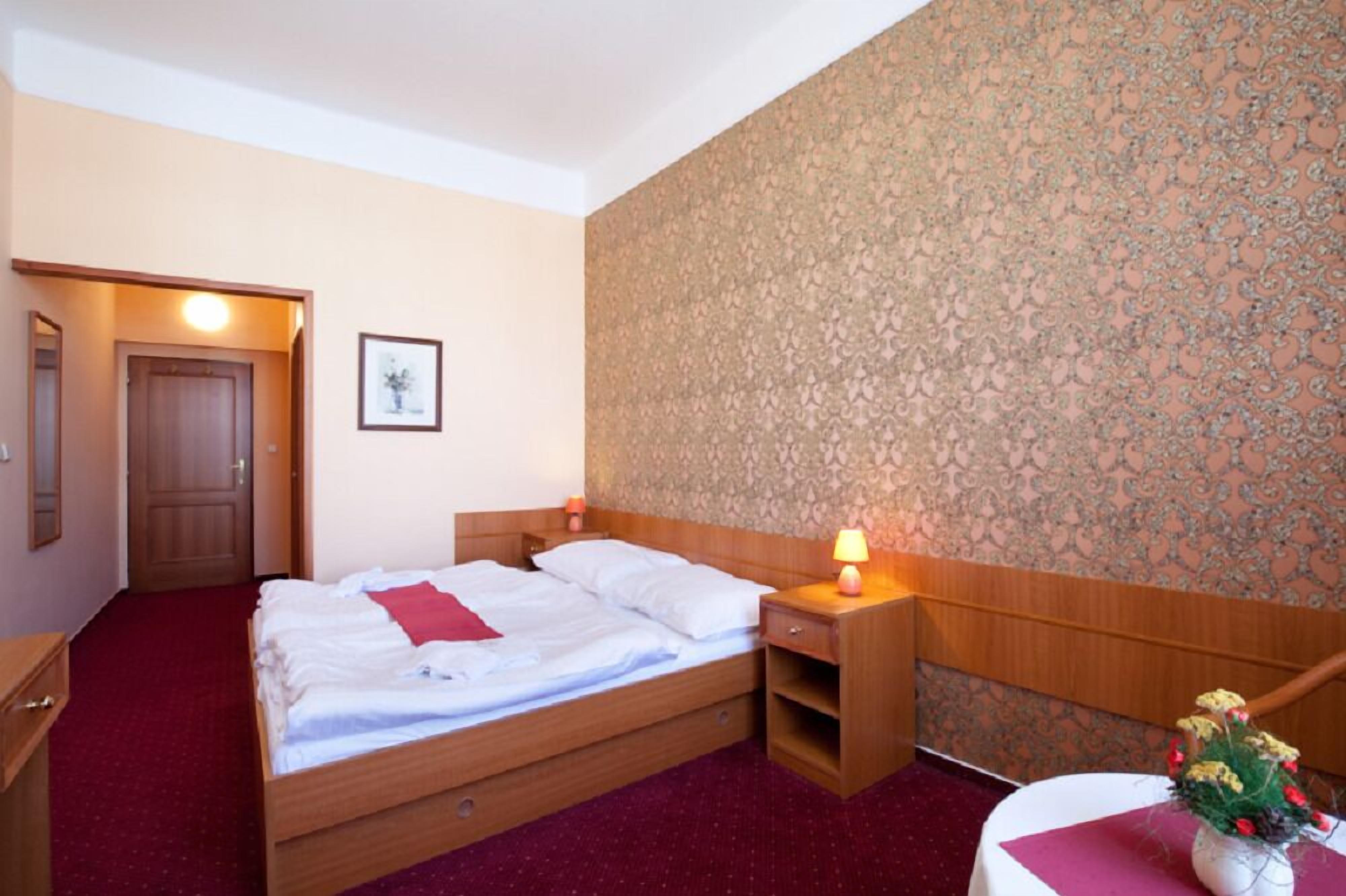 Quarto de hotel com cama de madeira, mesa de cabeceira com abajur, pequena mesa com um vaso de flores e porta que dá para outro cômodo.