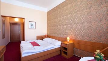 Quarto de hotel com cama de madeira, mesa de cabeceira com abajur, pequena mesa com um vaso de flores e porta que dá para outro cômodo.