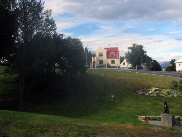 Un espacio cubierto de césped con una estatua, un edificio de techo rojo y una carretera al fondo.