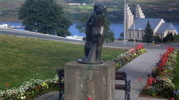 Escultura de una figura sobre un pedestal en una zona ajardinada con bancos y macizos de flores.