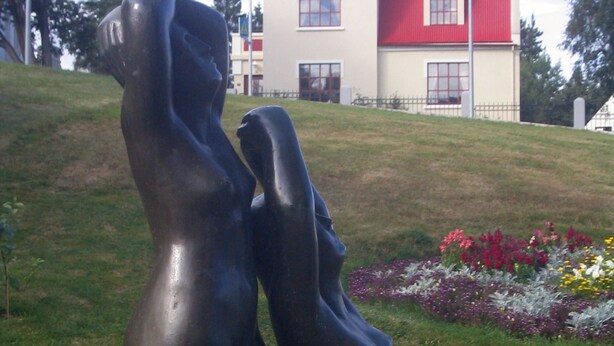 Une sculpture représentant deux personnages, placée sur un piédestal dans un espace verdoyant, avec un bâtiment en arri ère-plan.