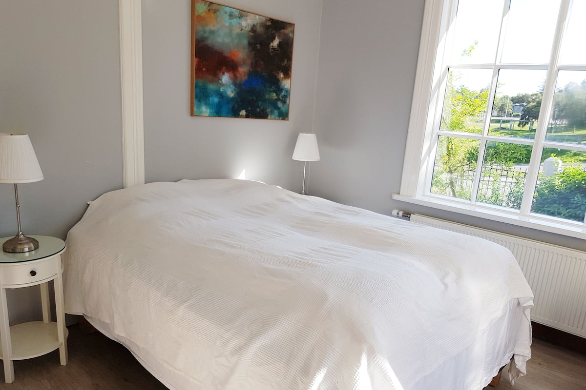 Un dormitorio con una cama grande, dos mesitas de noche con lámparas, una ventana con vistas a la vegetación y un cuadro abstracto y colorido en la pared.