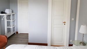 Une chambre d’hôtel avec un lit, une porte blanche, une table en verre avec une lampe, une étagère blanche avec des serviettes et une chaise.