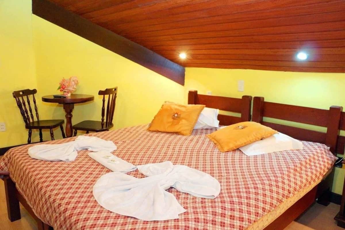 Une chambre avec une couvre-lit à carreaux, deux chaises en bois, une petite table ronde ornée d’un vase et un plafond en bois.