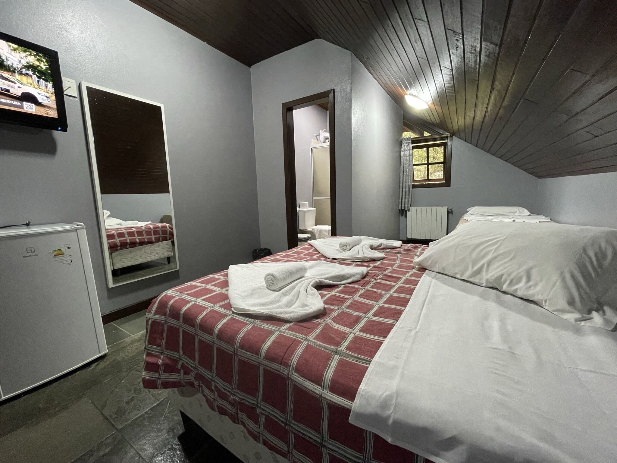A bedroom with a bed, a mirror, a small fridge, and a window.
