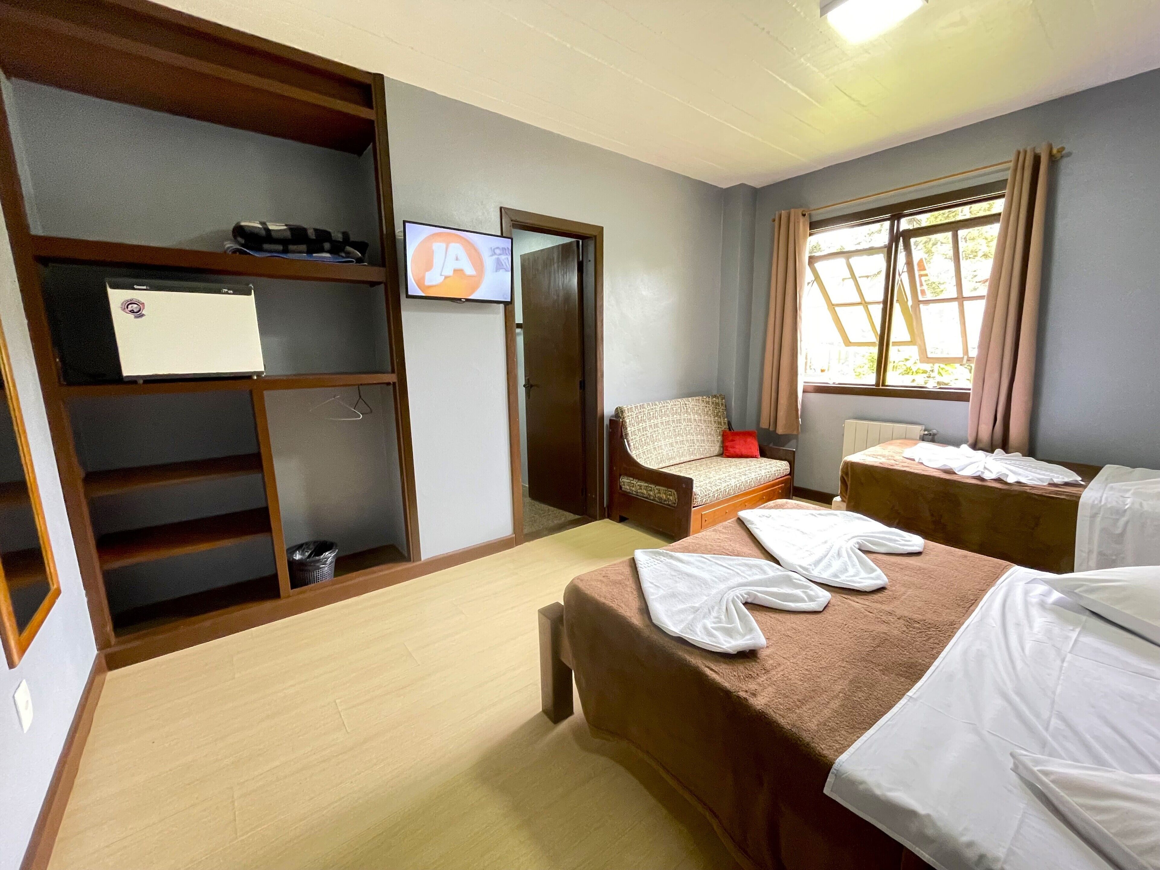 A hotel room with a wooden wardrobe, a sofa, two beds, a television, and a window with curtains.