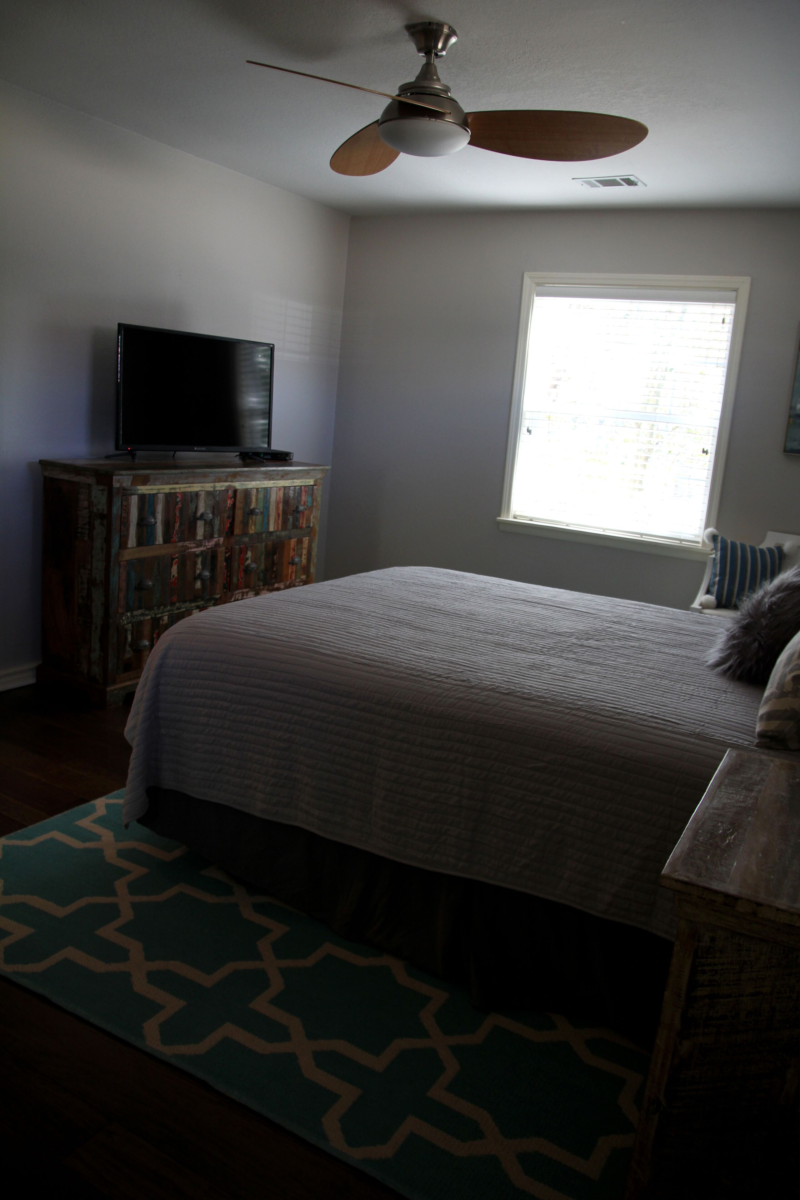A bedroom with a bed, a ceiling fan, a television on a wooden cabinet, and a window with blinds.