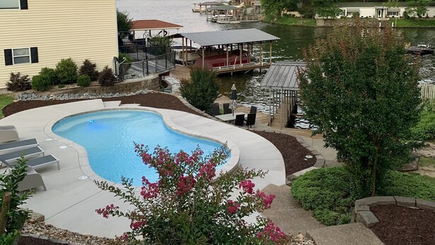A swimming pool with a curved edge, surrounded by a deck with lounge chairs. There is a dock with boats and a building with a green roof in the background.
