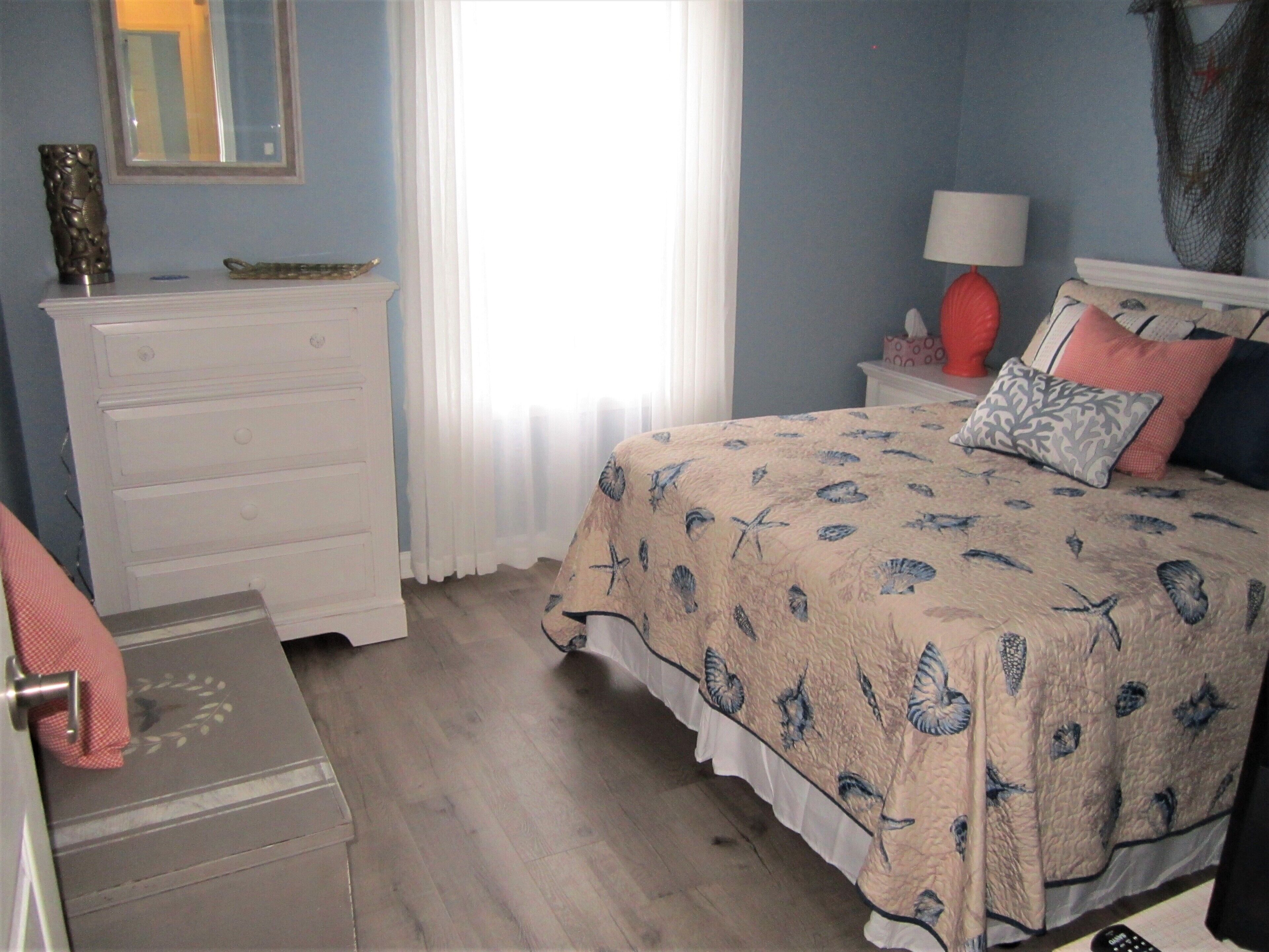 A bedroom with a bed, a dresser, a nightstand, a lamp, and a window with curtains.