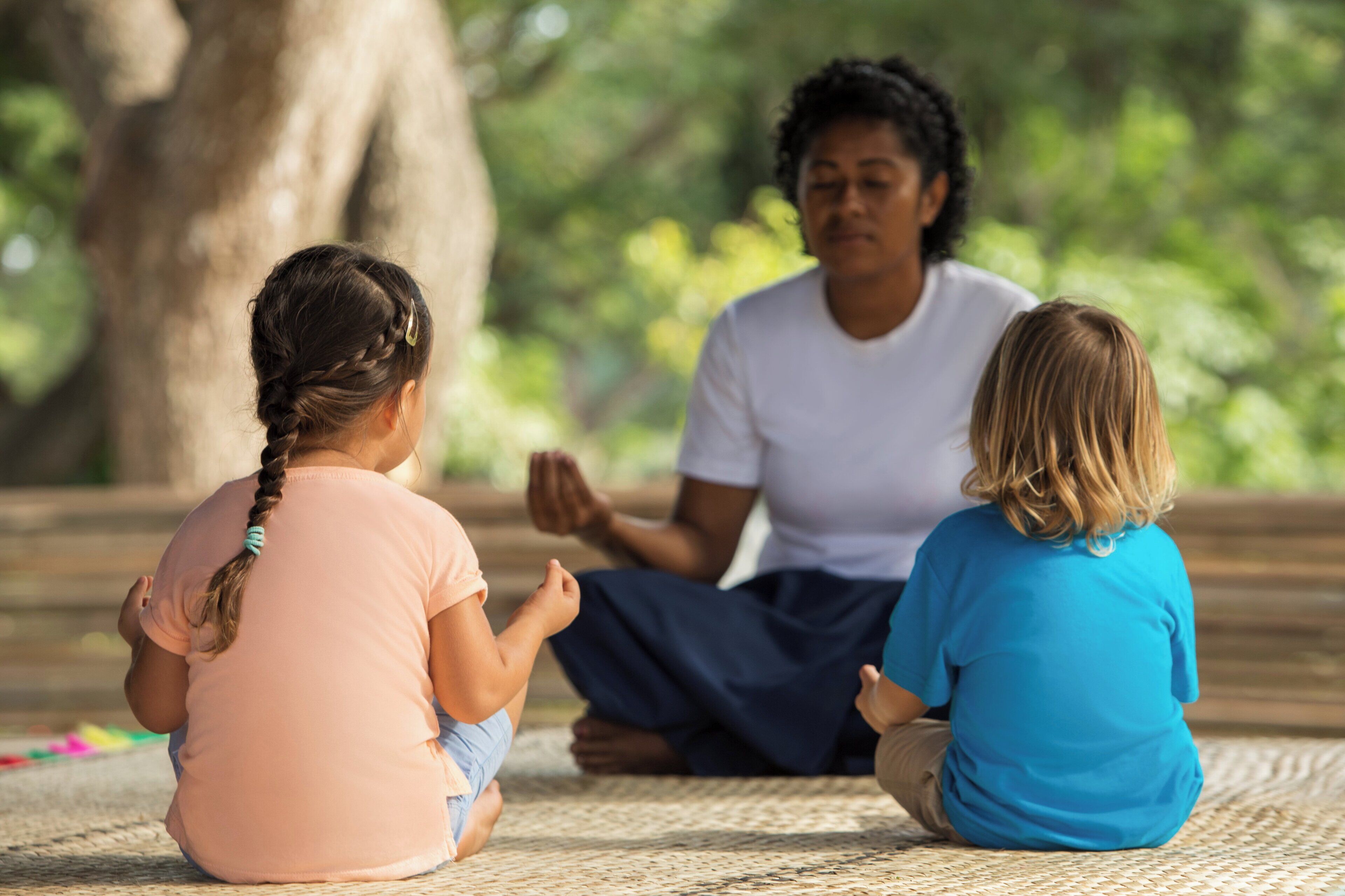Three individuals sitting on a woven mat outdoors, with a tree and greenery in the background.