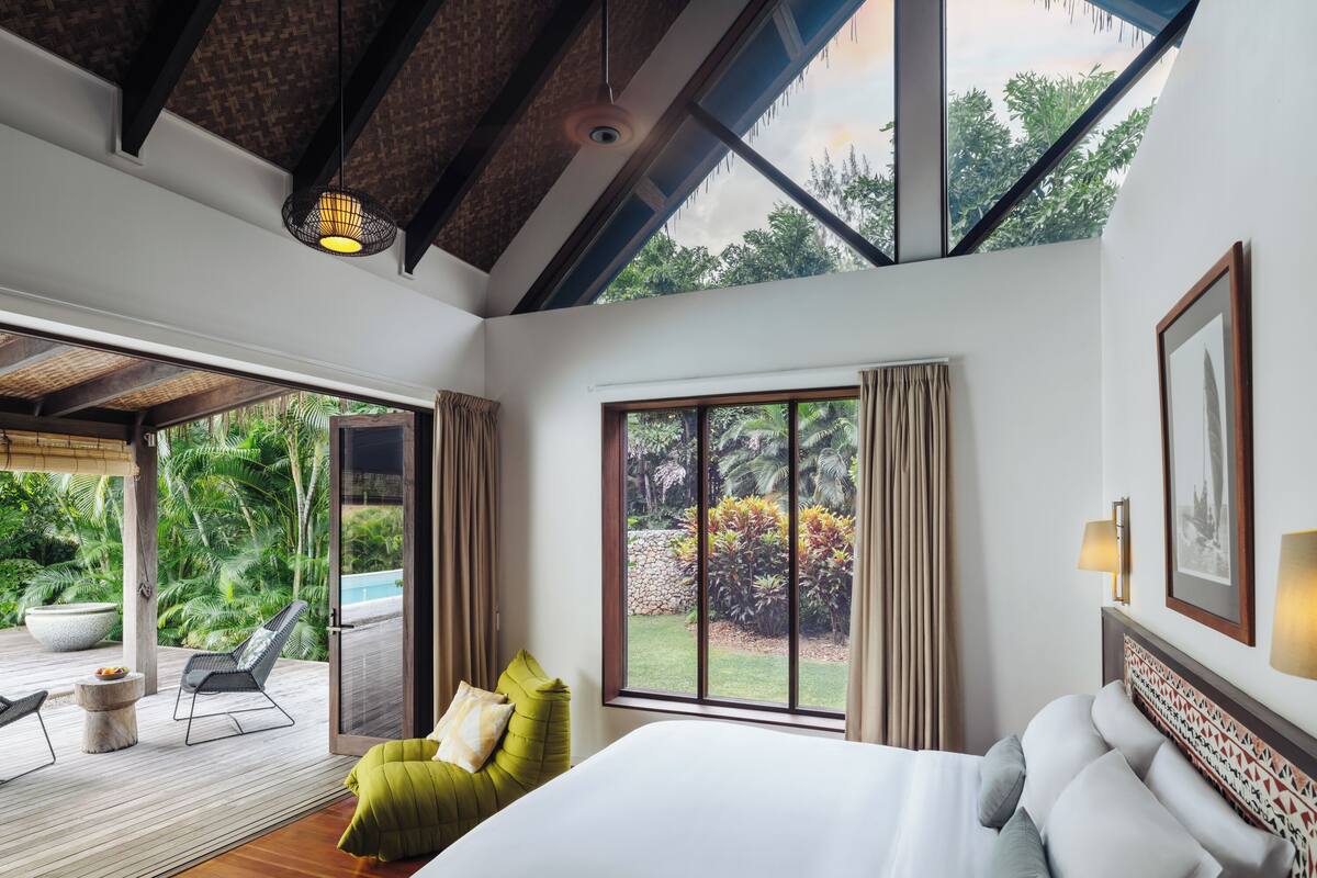 Un dormitorio con una cama grande, una silla verde y vistas a un jardín por un ventanal corredero.
