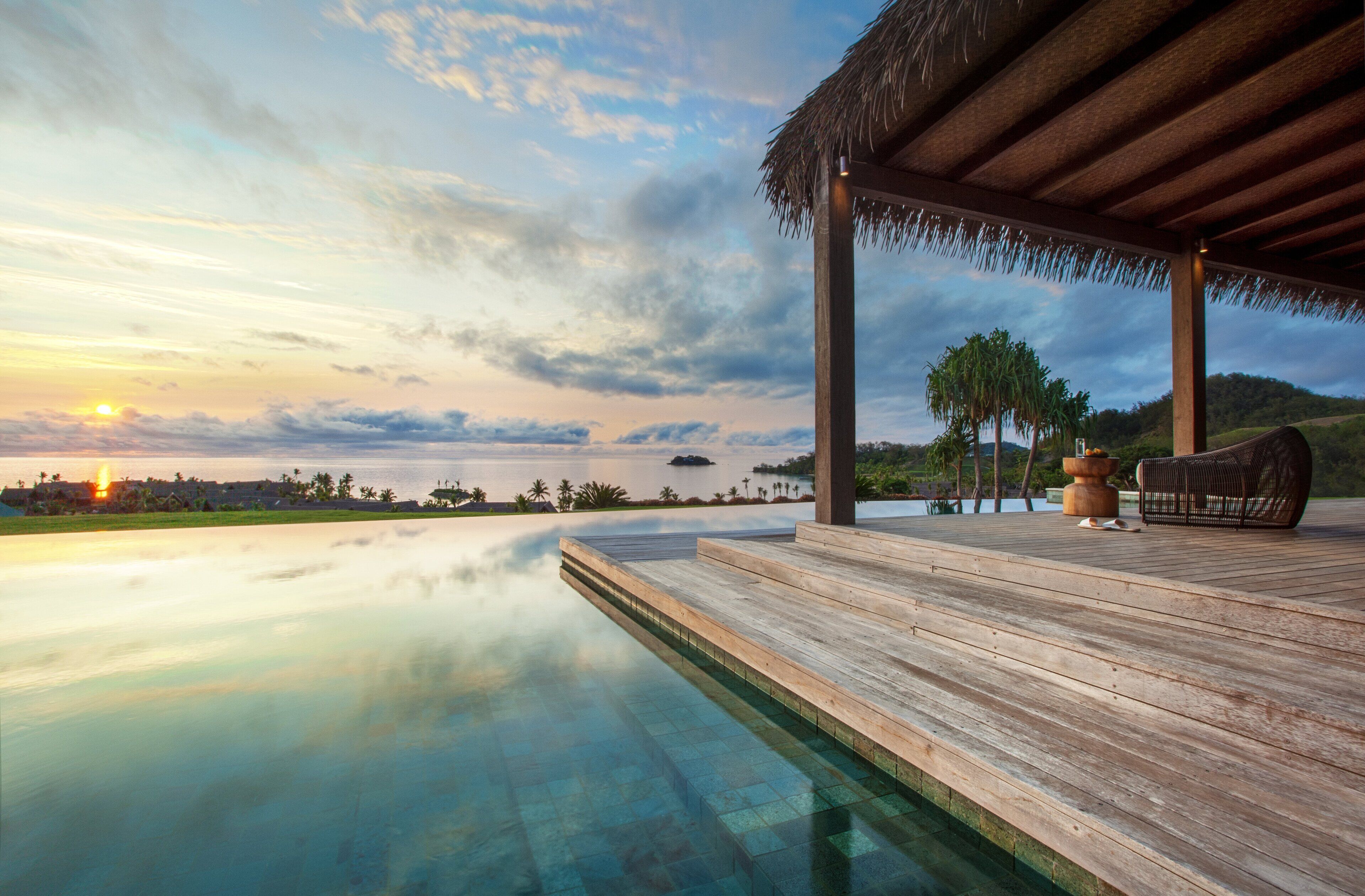 A wooden deck with a pool, a thatched roof structure, and a view of the sunset over the water.