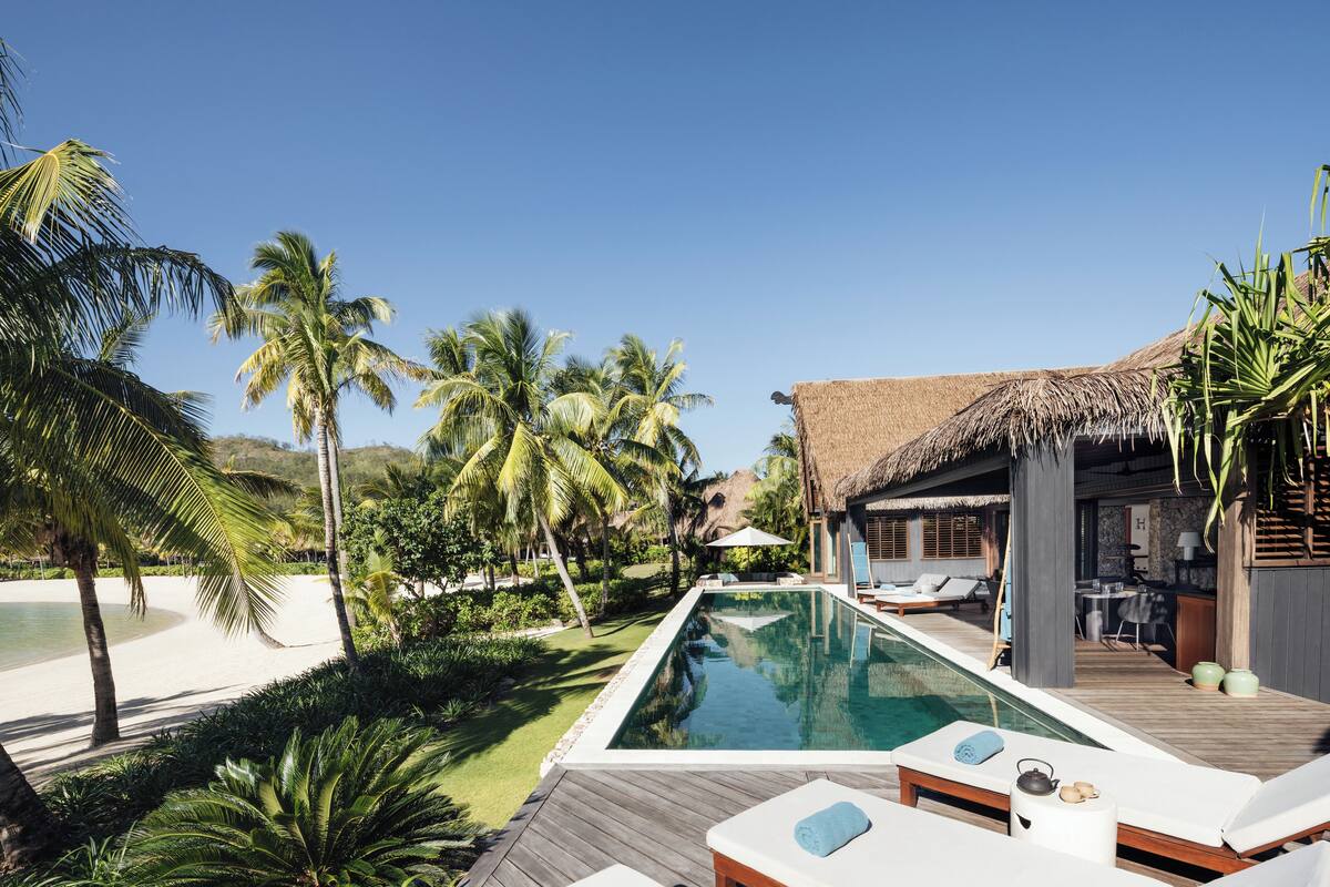 Zona junto a la piscina con sillones de descanso, una estructura de techo de paja y vistas a la playa y palmeras.