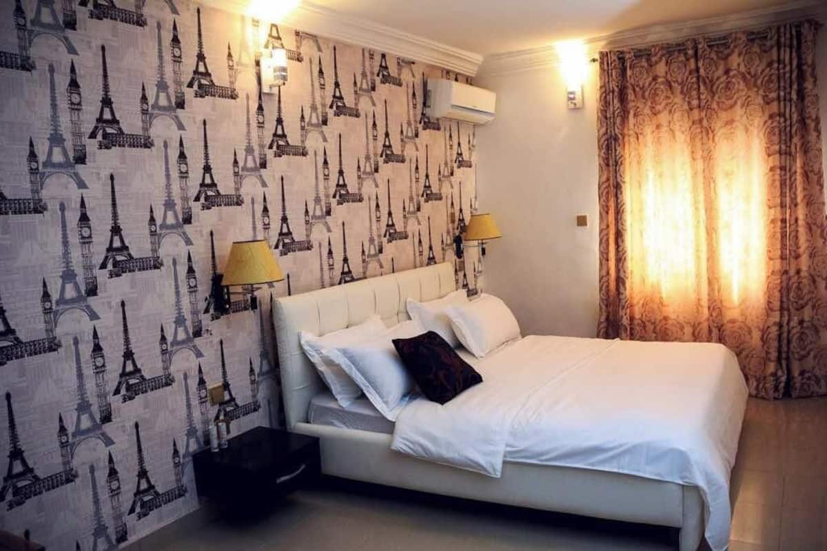 A hotel room with a bed, two bedside lamps, a patterned wallpaper, and a curtain.