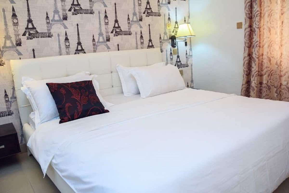 A hotel room with a bed, white bedding, a headboard with a patterned wallpaper, a bedside lamp, and a curtain.