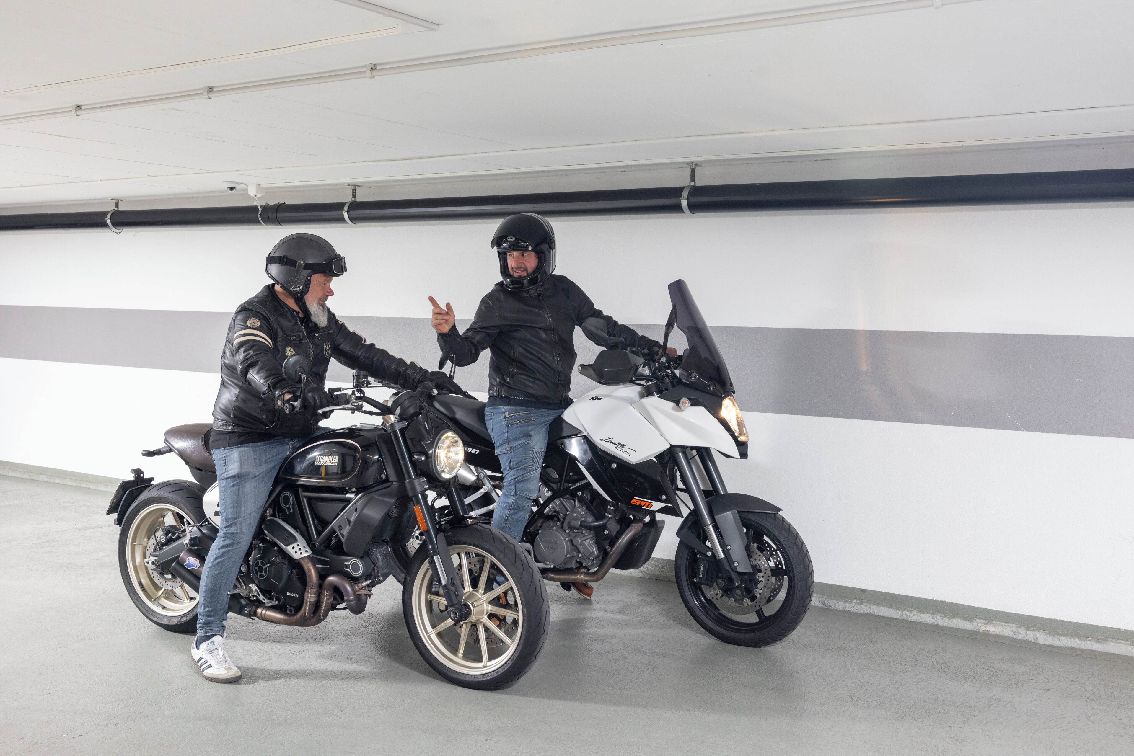 Dos motocicletas estacionadas en un garaje con una pared blanca y una banda negra.