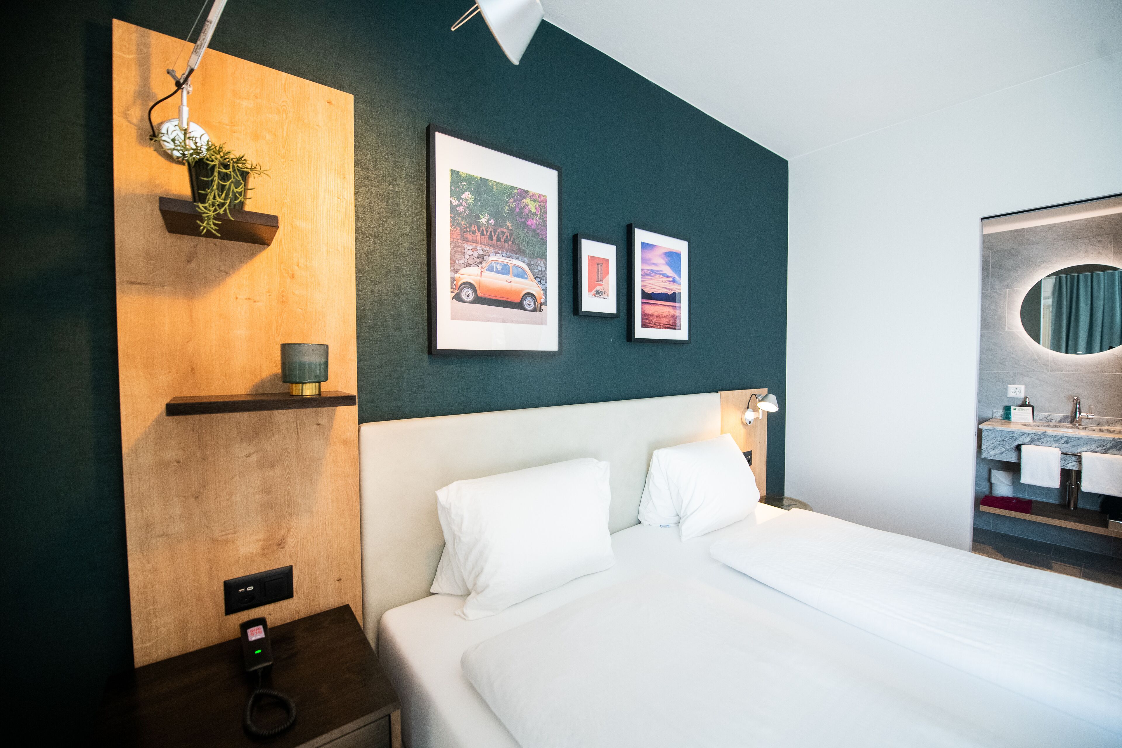 A modern hotel room with a bed, a bedside table with a lamp, a wall-mounted shelf with a plant, and framed pictures.