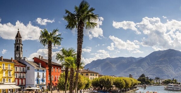 A coastal town with colorful buildings, a prominent clock tower, palm trees, and a mountain backdrop. - Ascona Lodge, Pool & Garden Retreat (Ascona)
