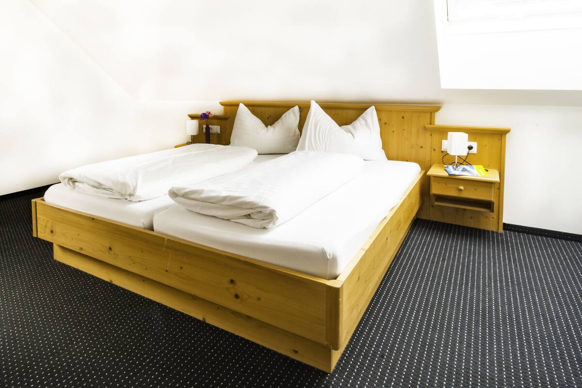 Un lit simple avec un cadre en bois et du linge de lit blanc, une table de chevet avec une lampe, et un sol à motifs en noir et blanc.